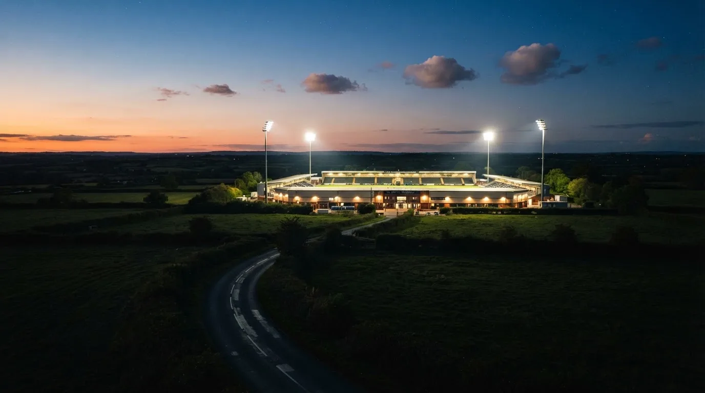 Stadion rozświetlony reflektorami widziany z daleka znad ciemnych pól w pogodny wieczór.