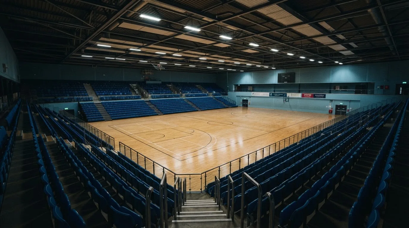 Duża hala sportowa widziana z daleka, parkiet jest pusty, a nad nim świecą rzędy reflektorów.