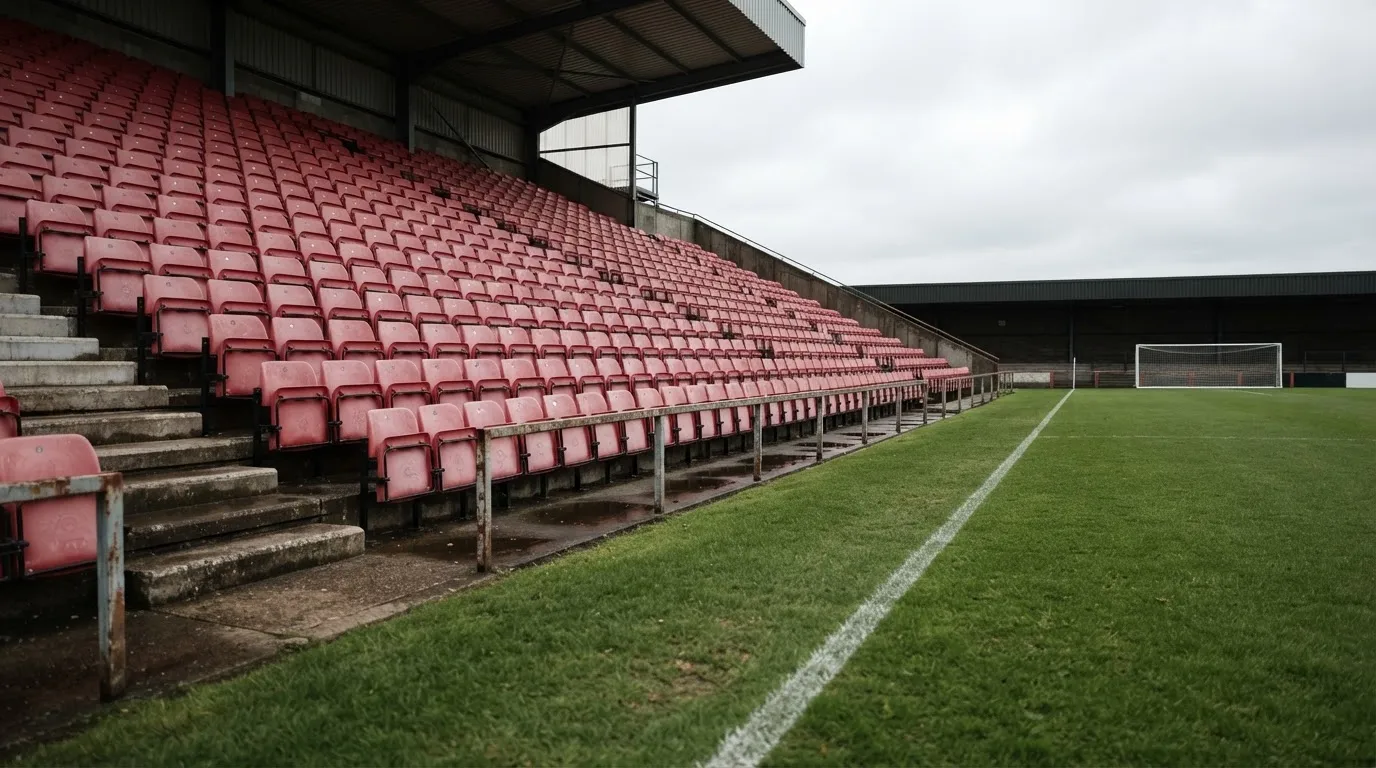 Rzędy pustych plastikowych krzesełek małego stadionu widziane z poziomu murawy pod pochmurnym niebem.