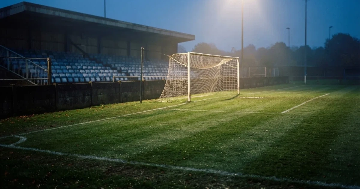 Pusta murawa stadionu miejskiego w chłodny jesienny wieczór, siatka bramki świeci w świetle reflektorów, nad boiskiem wisi mgła.