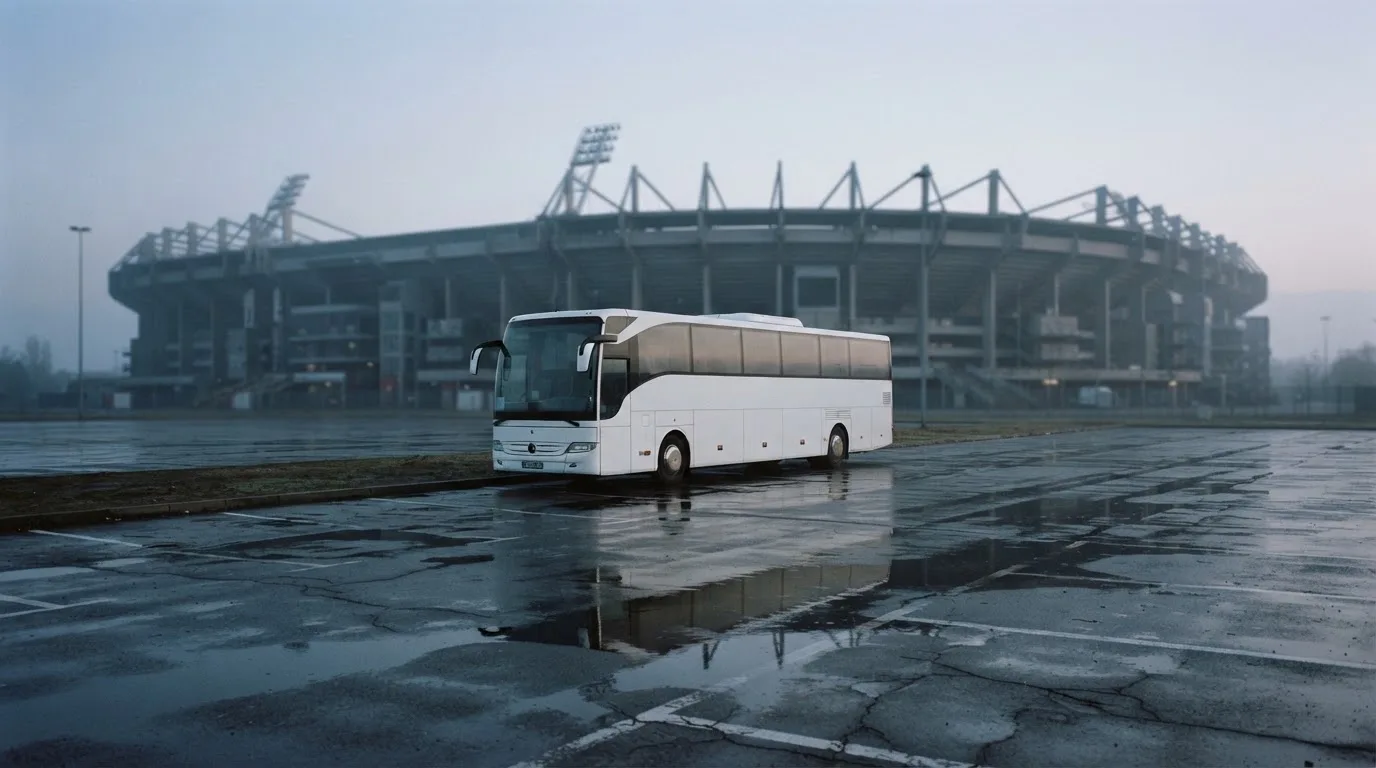 Autokar stoi na parkingu przed stadionem o świcie, rozmyte kontury trybun w tle, pusty plac i mokry asfalt.