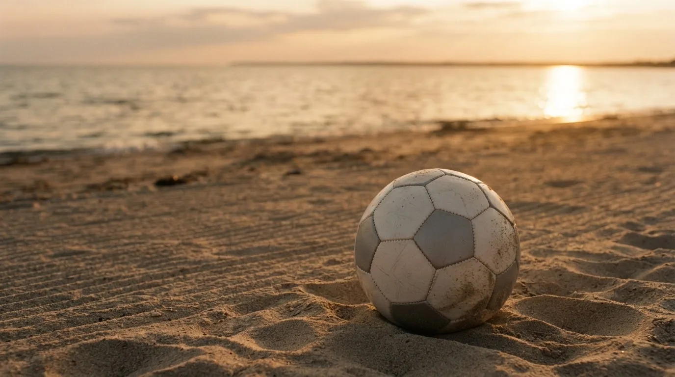 Piłka futbolowa leży na piasku boiska plażowego w ciepłym świetle zachodu, w tle morze i niski horyzont.