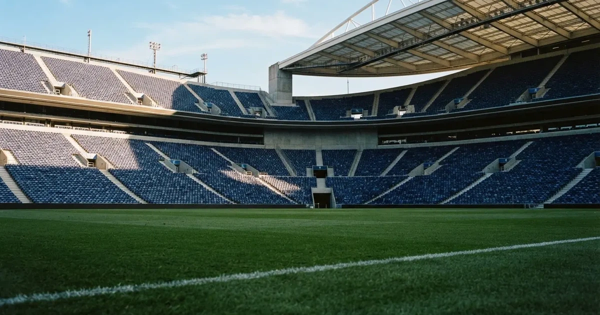 Trybuny wielkiego stadionu widziane z murawy, rzędy pustych ciemnoniebieskich krzesełek, ostre światło dnia i długie cienie.