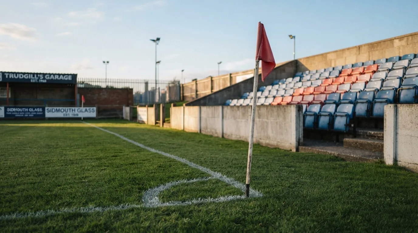 Chorągiewka rożna wbita w trawę na skraju boiska, za nią rzędy pustych plastikowych krzesełek na trybunie.