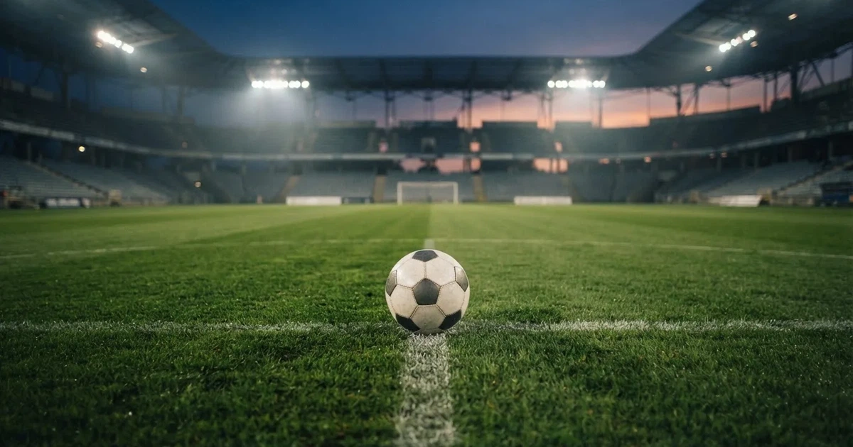 Piłka nożna leży na środku murawy dużego stadionu o zmierzchu, a nad boiskiem świecą rzędy reflektorów nad pustymi trybunami.