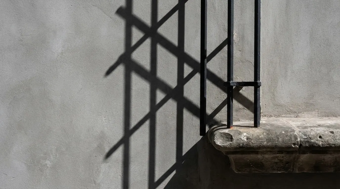 Cień okiennej kraty pada na szarą ścianę, obok widać fragment ciężkiego parapetu.